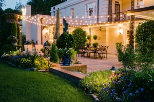 Patio with outdoor string lights in between a tidy lawn and the back of a large house.